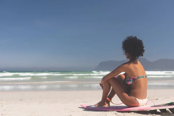 侧视图的混合种族女性冲浪者与冲浪板坐在海滩上 她在看大海 — 图库照片