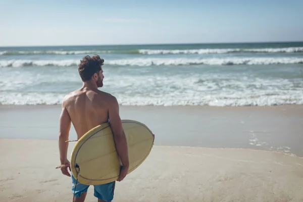 在阳光明媚的日子里 年轻的白人男性冲浪者的后视 冲浪板站在海滩上 他在看海浪 — 图库照片