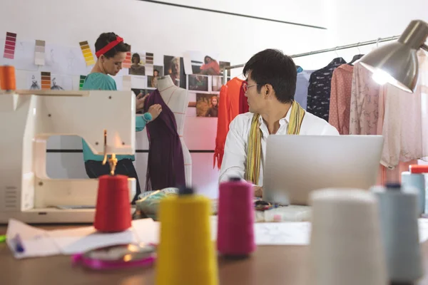 デザインスタジオでラップトップで作業しながら ミックスレースの女性デザイナーと話をするアジアの男性ファッションデザイナーの正面図 — ストック写真