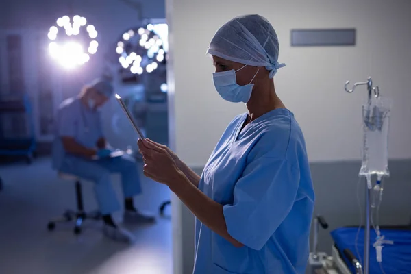 男性が病院で彼女の後ろの手術室に書いている間 手術室でデジタルタブレットを使用している白人女性外科医の側面図 — ストック写真