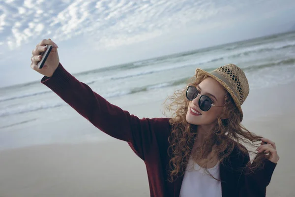 帽子とサングラスのビーチで Selfie 彼女の携帯電話の立っているを取って美しい若い白人女性の高角度のビュー 彼女は笑っています — ストック写真