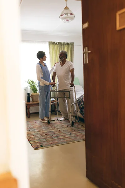 老人ホームで歩行者と歩きながら アフリカ系アメリカ人男性と話をしているアジアの女性看護婦の正面図 — ストック写真