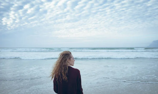 美丽的年轻高加索妇女与卷发站在面对大海在海滩上的后面的看法 她把目光移开 — 图库照片