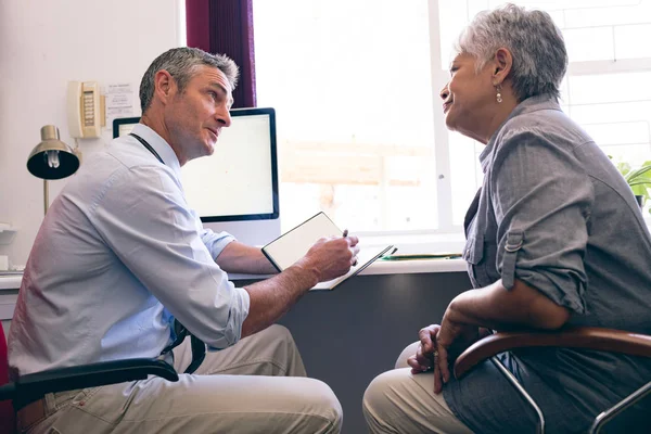 Side view of male Caucasian doctor prescribing medicine to senior African American woman in clinic at retirement home