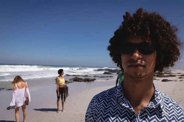 背景に彼の友人と太陽の下でビーチに立っている間 カメラを見て若いミックスレースの男の正面図 — ストック写真