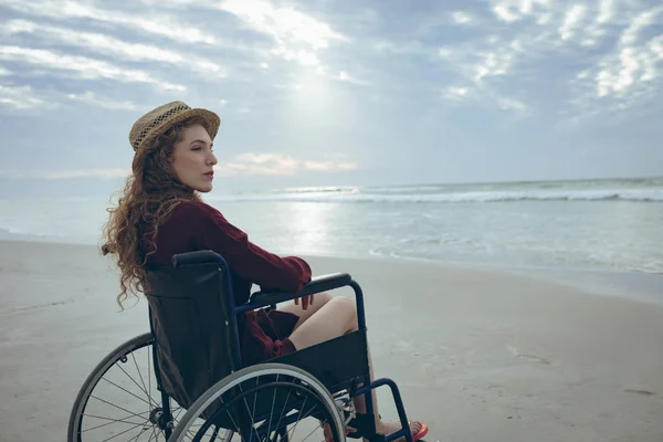 年轻的残疾白人妇女坐在海滩上的轮椅侧视图 她把目光移开 — 图库照片