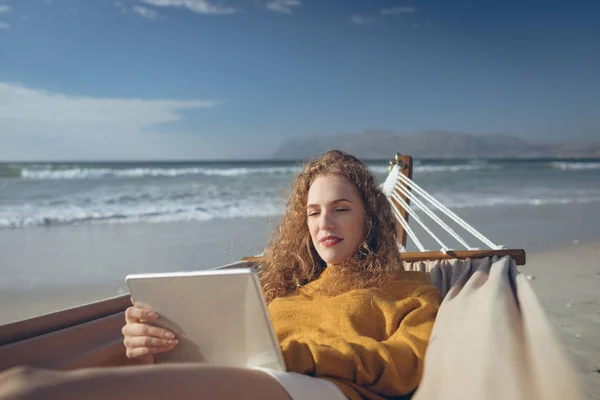 晴れた日にビーチでハンモックに横たわっている間デジタル タブレットを使用して美しい白人女性の正面図 — ストック写真
