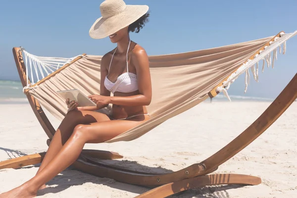 在阳光明媚的日子里 穿着比基尼的年轻混血女子坐在沙滩上的吊床上 用数字平板电脑戴着帽子 可以看到前厅的景色 — 图库照片