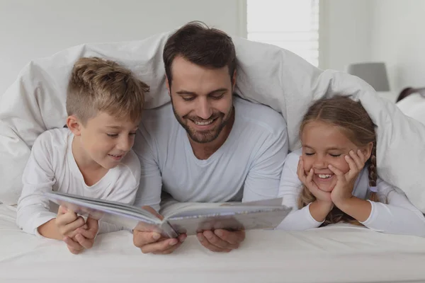 在舒适的家中 快乐的白种人父亲和孩子在毯子下看书的前视图 — 图库照片
