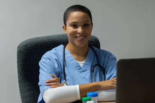 病院の診察室の机に座りながらカメラを見ながら腕を組んだ幸せな混血女子医師の正面図 — ストック写真