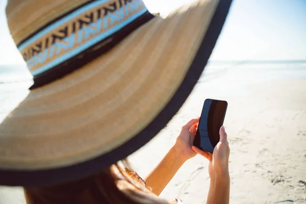 太陽の下でビーチで携帯電話を使用して帽子をかぶった女性の側面図 — ストック写真