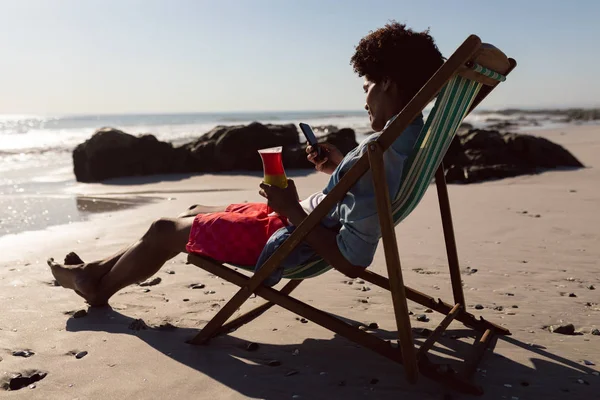 ビーチチェアでカクテルを飲みながら携帯電話を使ったアフリカ系アメリカ人の若者の側面図 — ストック写真