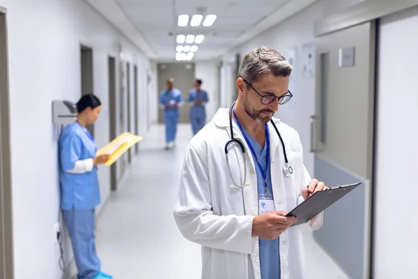 Vista Frontal Del Doctor Varón Caucásico Escribiendo Portapapeles Pasillo Hospital — Foto de Stock