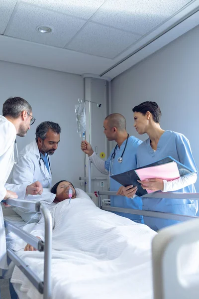 アジアの女性患者が病院でベッドに横たわっている間 多様な医師がIv点滴と酸素マスクを調整する側面図 — ストック写真