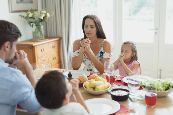 快適な家のダイニングテーブルで昼食をとる前に一緒に祈る白人家族の正面図 — ストック写真