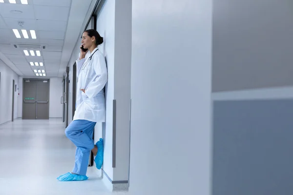 病院で壁に寄りかかっている間に携帯電話で話す白人女性医師の側面図 — ストック写真