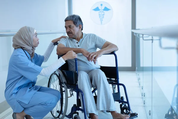 病院の廊下で車椅子で障害のあるシニア混合レース男性患者と相互作用するヒジャーブの混合人種女性医師の側面図 — ストック写真