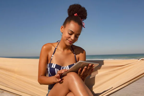 太陽の下でビーチでハンモックに座っている間 デジタルタブレットを使用して美しいアフリカ系アメリカ人女性の正面図 — ストック写真