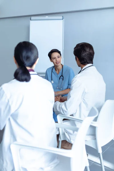 病院の会議室のテーブルで座って話し合う多様な医療チームと話す女性白人看護師の正面図 — ストック写真
