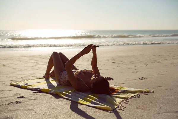 白种妇女使用手机的侧视图 而放松在野餐毯在海滩在阳光下 — 图库照片