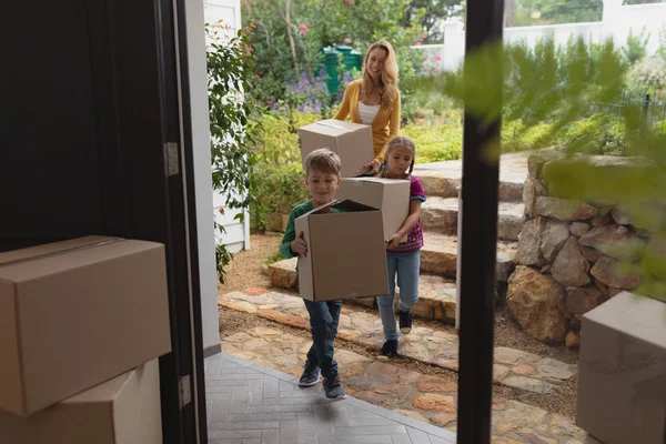 ダンボール箱を持つ白人の母と子供が家に向かって歩いている正面図 — ストック写真