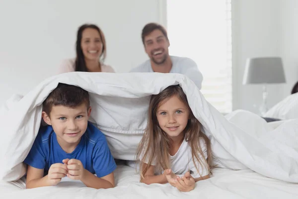 自宅の寝室でベッドに座っている親の間に毛布の下に横たわっている白人の子供の肖像画 — ストック写真