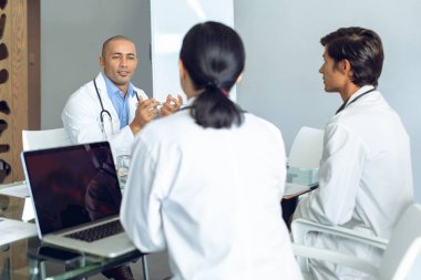 Front view of male Mixed-race doctor attentive to diverse medical team sitting and discussing at the table of conference room in hospital clipart