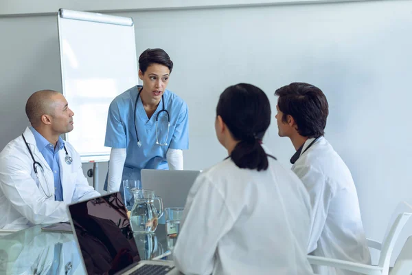 病院の会議室のテーブルに座って気配りの多様な医療チームに話す立っている女性白人看護師の正面図 — ストック写真
