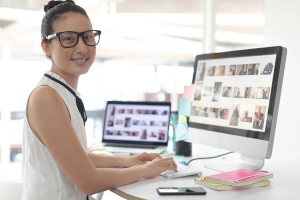 現代のオフィスで机の上でコンピュータで作業しながらカメラを見ているアジアの女性グラフィックデザイナーの正面図 — ストック写真