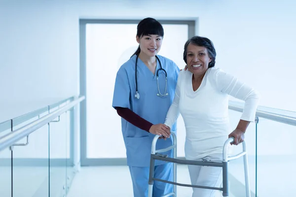 首の周りに聴診器を持つアジアの女性医師の正面図は 成熟した混合人種の女性患者が病院の廊下で歩行者と一緒に歩くのを助ける — ストック写真