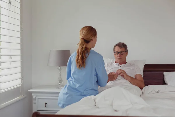 Rear view of Caucasian female doctor talking to disabled active senior man about his hearing aid in bed in bedroom at home