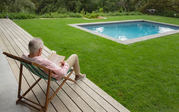自宅の裏庭でサンラウンジャーでリラックスしながら本を読む現役の白人男性の背面図 — ストック写真