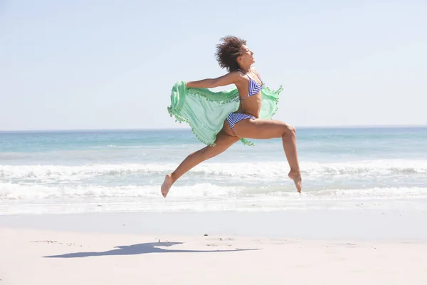身穿比基尼 头戴围巾跳到海滩上的非洲裔美国妇女的侧面照片 — 图库照片