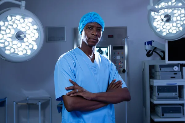 病院の手術室に立って腕を組んだ深刻なアフリカ系アメリカ人男性外科医の正面図 — ストック写真