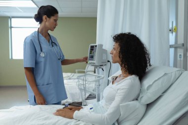 Side view of mixed race female doctor adjusting medical monitor while mixed race female patient sitting on bed in the ward at hospital clipart