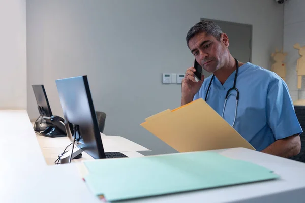 白种男性外科医生在医院办公桌前查看医疗报告时用手机交谈的侧面视图 — 图库照片