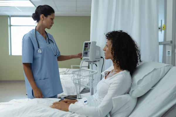 病院の病棟のベッドに座っている混合レース女性患者が医療モニターを調整する混合人種女性医師の側面図 — ストック写真