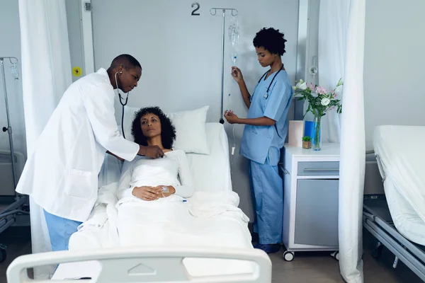 在医院的病房里 非洲裔美国男医生用听诊器检查混血女性患者的正面观点 旁边非洲裔美国女医生调整Iv滴注 — 图库照片