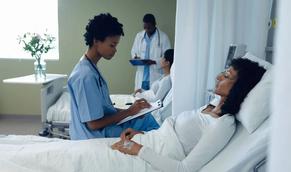 病院の病棟のクリップボードに書き込むアフリカ系アメリカ人女性医師の側面図 背景に混合人種女性患者とアフリカ系アメリカ人の男性医師が話す — ストック写真