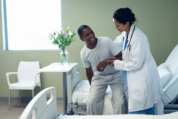 アフリカ系アメリカ人男性患者が病院の病棟の病院のベッドに立ち上がるのを助ける混合人種女性医師の側面図 — ストック写真