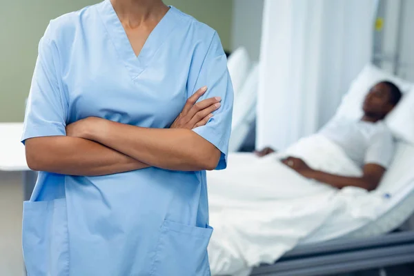 混血女医生站在手臂交叉 而非洲裔美国人睡在医院的病房的床上 — 图库照片