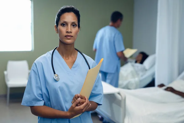 病院の病棟でベッドに横たわる白人女性と話し合いながら ファイルと共に立つ混合人種女性医師の肖像 — ストック写真