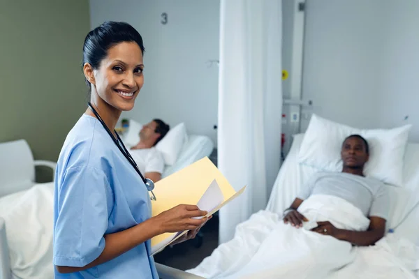 混血女医生在看医疗报告 而两名病人男子躺在医院病房的病床上的肖像 — 图库照片