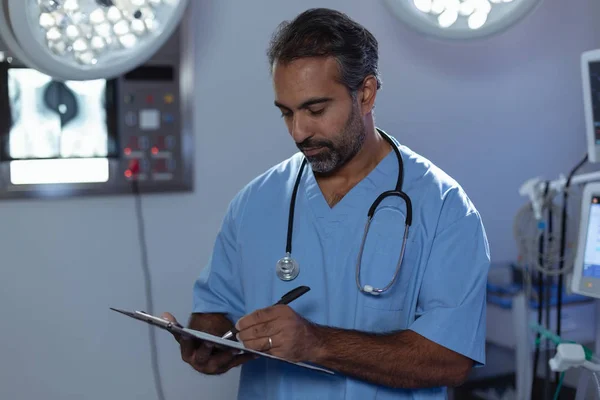 Vooraanzicht Van Volwassen Mixed Race Mannelijke Chirurg Schrijven Klembord Operatiekamer — Stockfoto