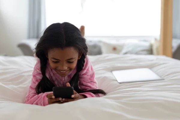 自宅の寝室のベッドで携帯電話を使用してアフリカ系アメリカ人の女の子の正面図 — ストック写真
