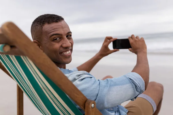 Portret Afroamerykańskiego Człowieka Klikając Zdjęcia Telefonem Komórkowym Siedząc Krześle Plażowym — Zdjęcie stockowe