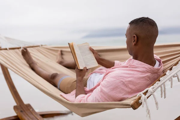 Bakifrån Afroamerikanska Mannen Läser Bok Samtidigt Koppla Hängmatta Stranden — Stockfoto