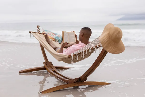 Tylny Widok Afroamerykanina Czytającego Książkę Podczas Relaksu Hamaku Plaży — Zdjęcie stockowe
