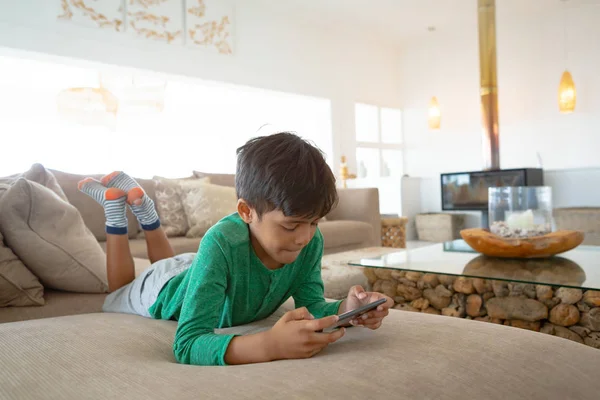 自宅のソファに横たわって携帯電話でゲームをしているかわいい混合レースの少年の正面図 — ストック写真