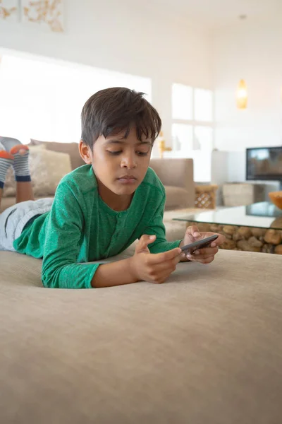 自宅のソファに横たわって携帯電話でゲームをしている若いかわいい混合レースの少年の正面ビュークローズアップ — ストック写真